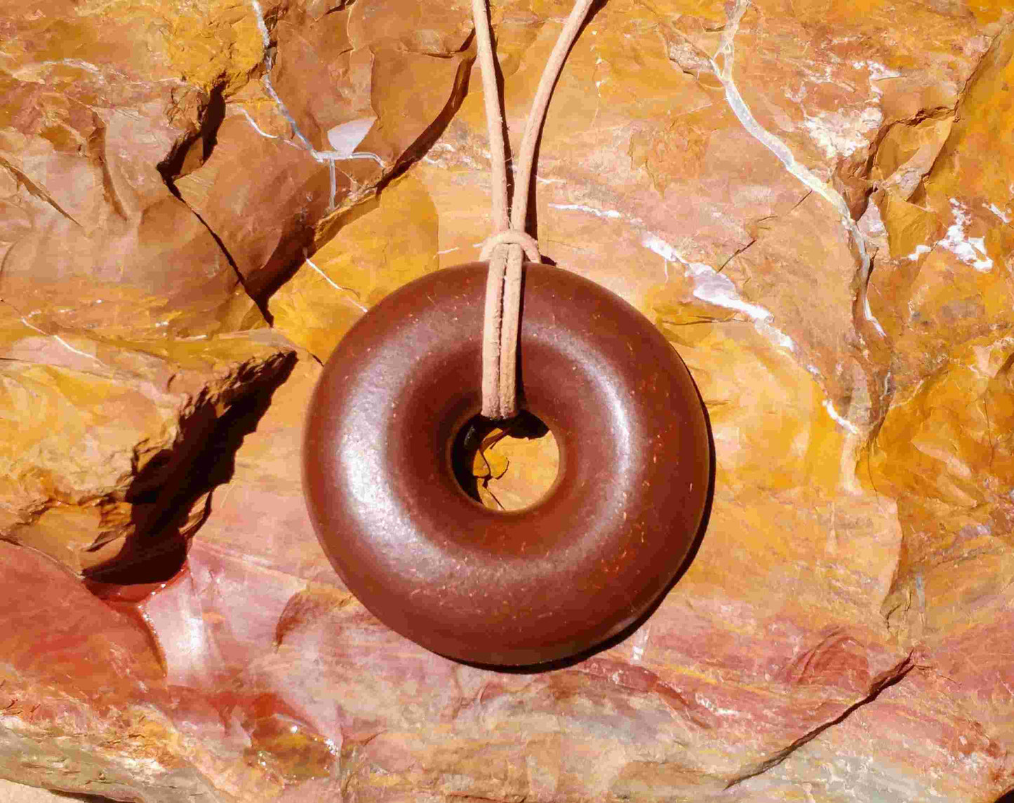 A brown donut-shaped Orgonite pendant with a leather necklace cord, placed against a background of red rocks. andy orgone emf sedona vortex jewlery red jasper