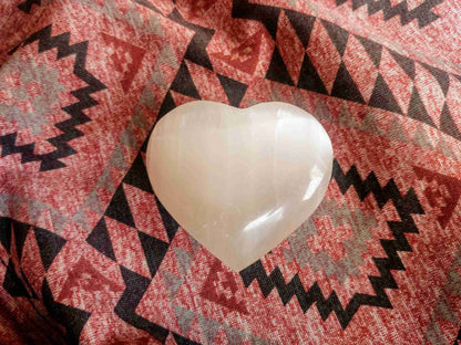 White Selenite Puffy Heart – Reiki Healing Crystal & Energy Cleanser displayed on a red geometric fabric background. Ideal for meditation and energy work.